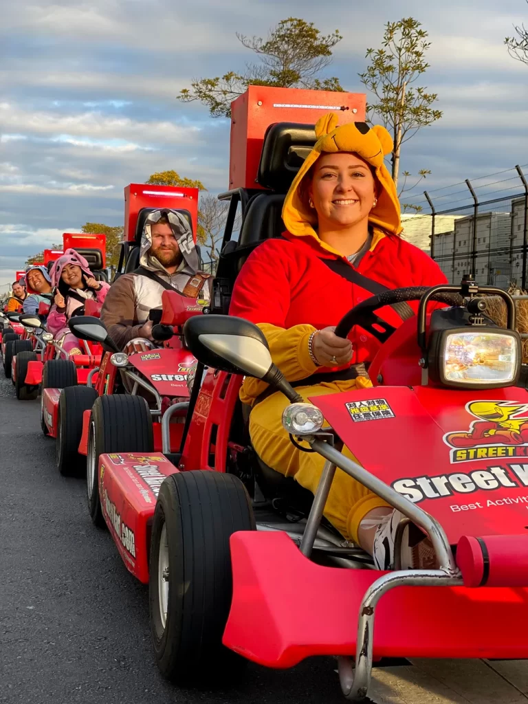 IMG_1123 - StreetKart Woman in a yellow bear hoodie drives a red go-kart, smiling, with a line of riders in costumes behind her.
