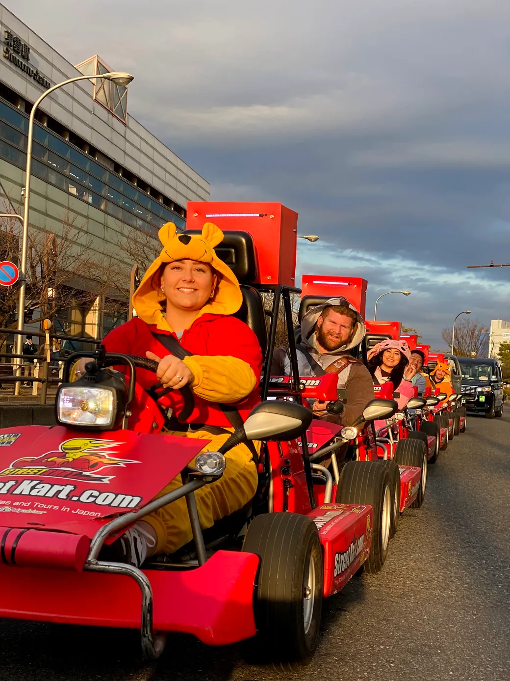 Smiling woman in a yellow animal hood drives a red go-kart at a street parade, with more costumed riders in red karts behind her.