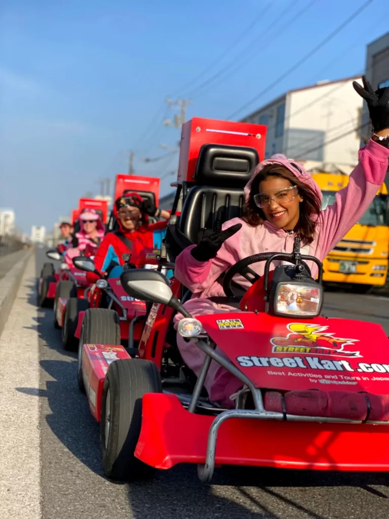 IMG_4093 - StreetKart Smiling person in a pink hoodie drives a red go-kart and waves, part of a line of pink-clad riders in a city street parade.