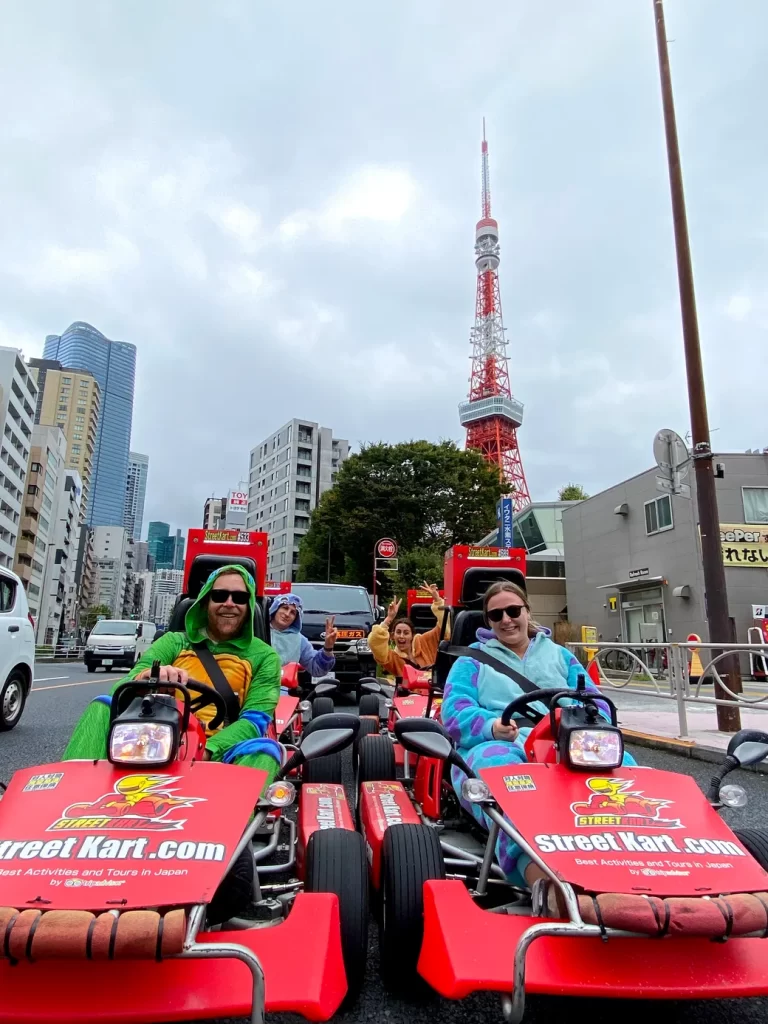 IMG_6165 - StreetKart Group of people in red street go-karts on a city street with Tokyo Tower in the background, wearing colorful outfits.
